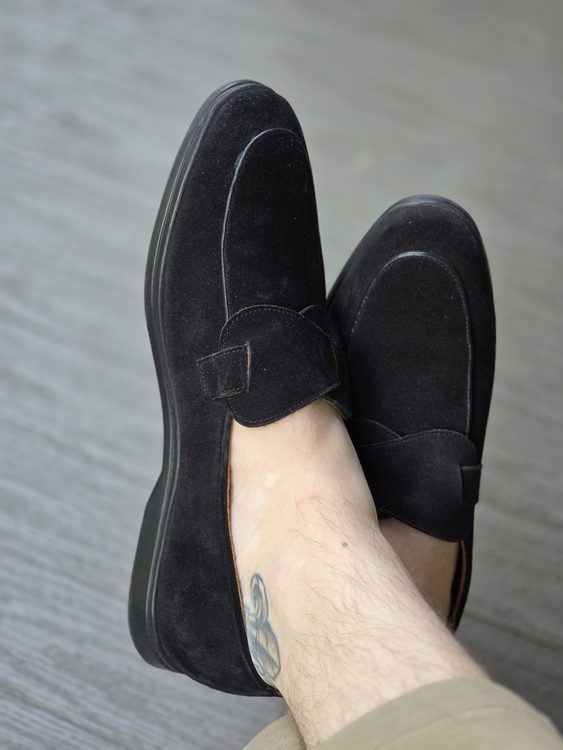 Black suede shoes with a decorative strap, worn on a foot with a visible tattoo.