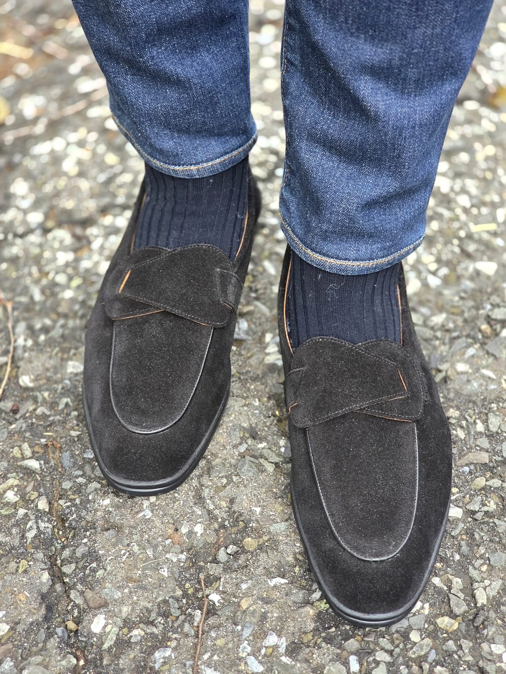 Black suede shoes with a decorative strap, paired with dark ribbed socks and blue denim jeans.
