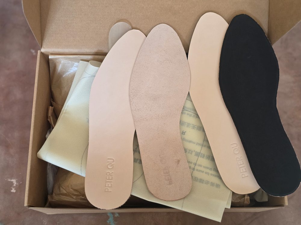 A box contains various shoe insoles in light beige, dark black, and a textured green material, with "PETER QU" embossed on the beige insoles.