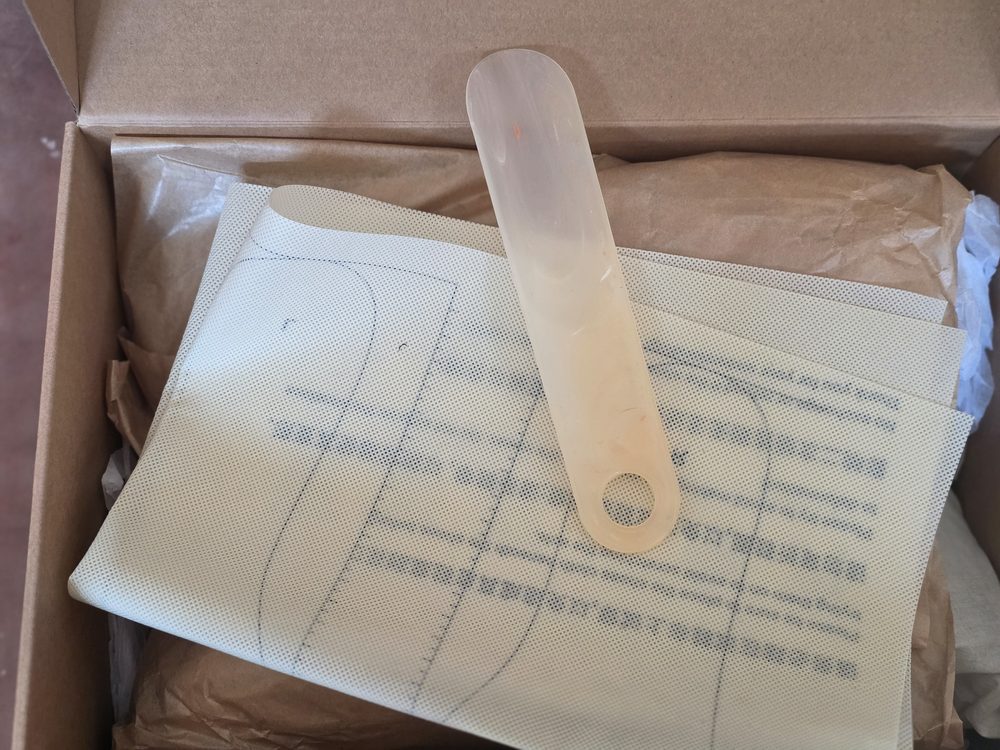 A light-colored, perforated leather piece is displayed alongside a transparent plastic tool inside a cardboard box.