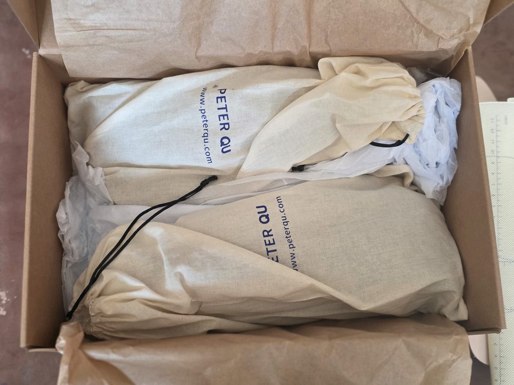 Two beige fabric bags with black drawstrings are neatly packed inside a cardboard box, labeled with "PETER QU."