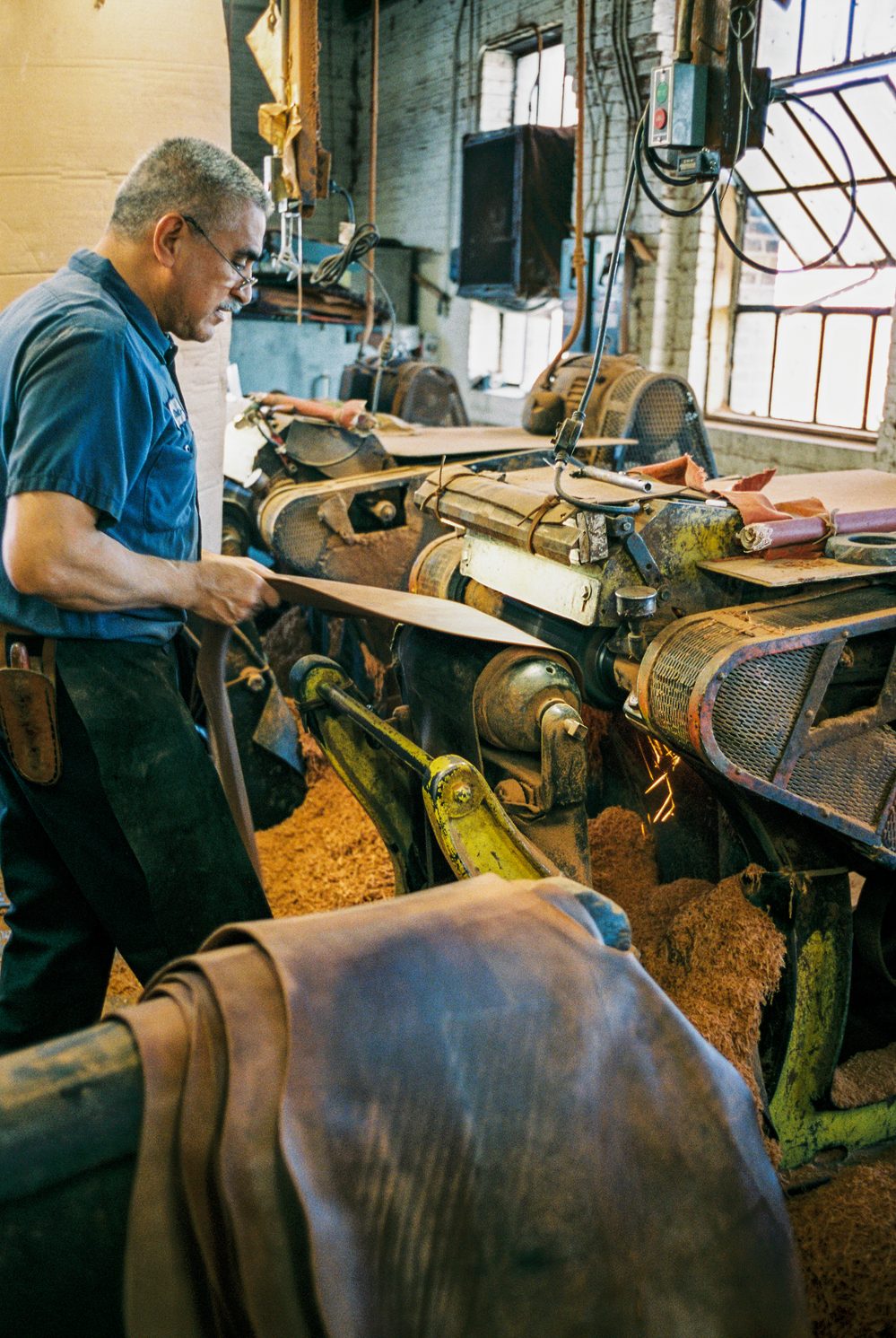 A craftsman in a blue shirt operates a leather cutting machine, showcasing materials for the Carmina Bespoke Trunk Show.