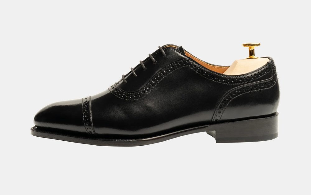 A black leather dress shoe featuring brogue detailing and a wooden shoe tree.