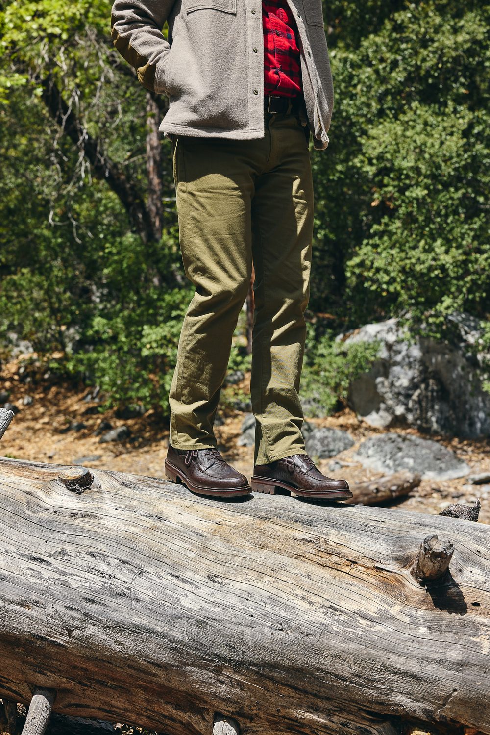 A person stands on a log wearing brown leather shoes, olive green pants, and a gray jacket over a red flannel shirt.