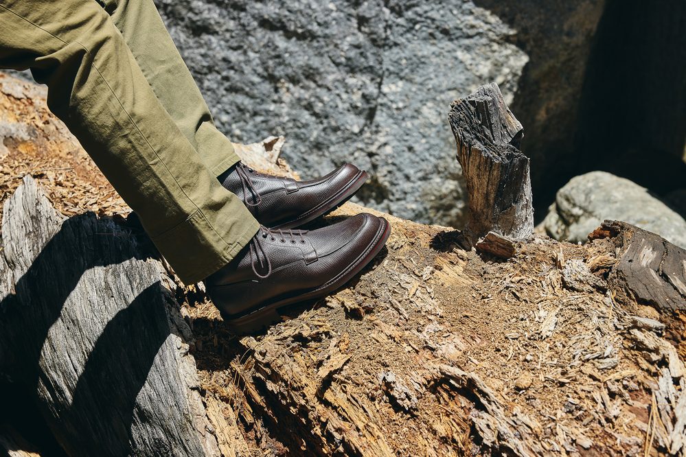 A close-up of dark brown leather shoes resting on a log, paired with olive green pants.