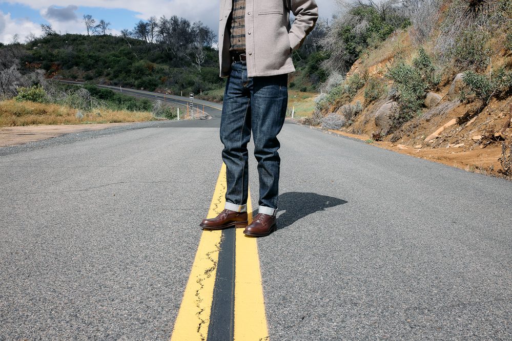 A person stands on a road wearing dark denim jeans and brown leather shoes, with a light-colored jacket.