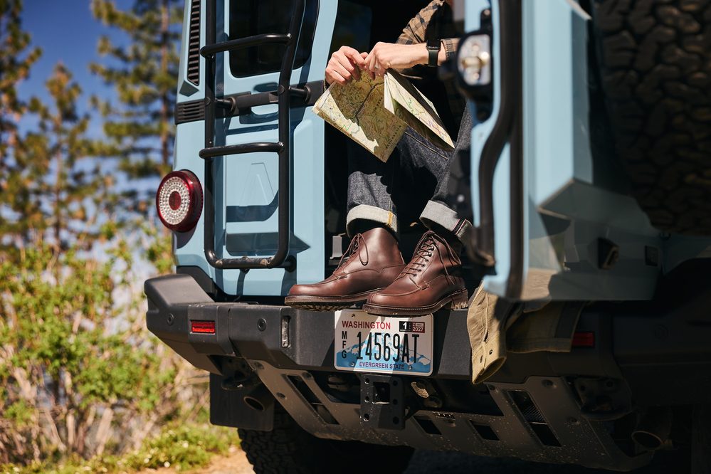 A person wearing brown leather boots sits on the back of a blue vehicle, holding a folded map.