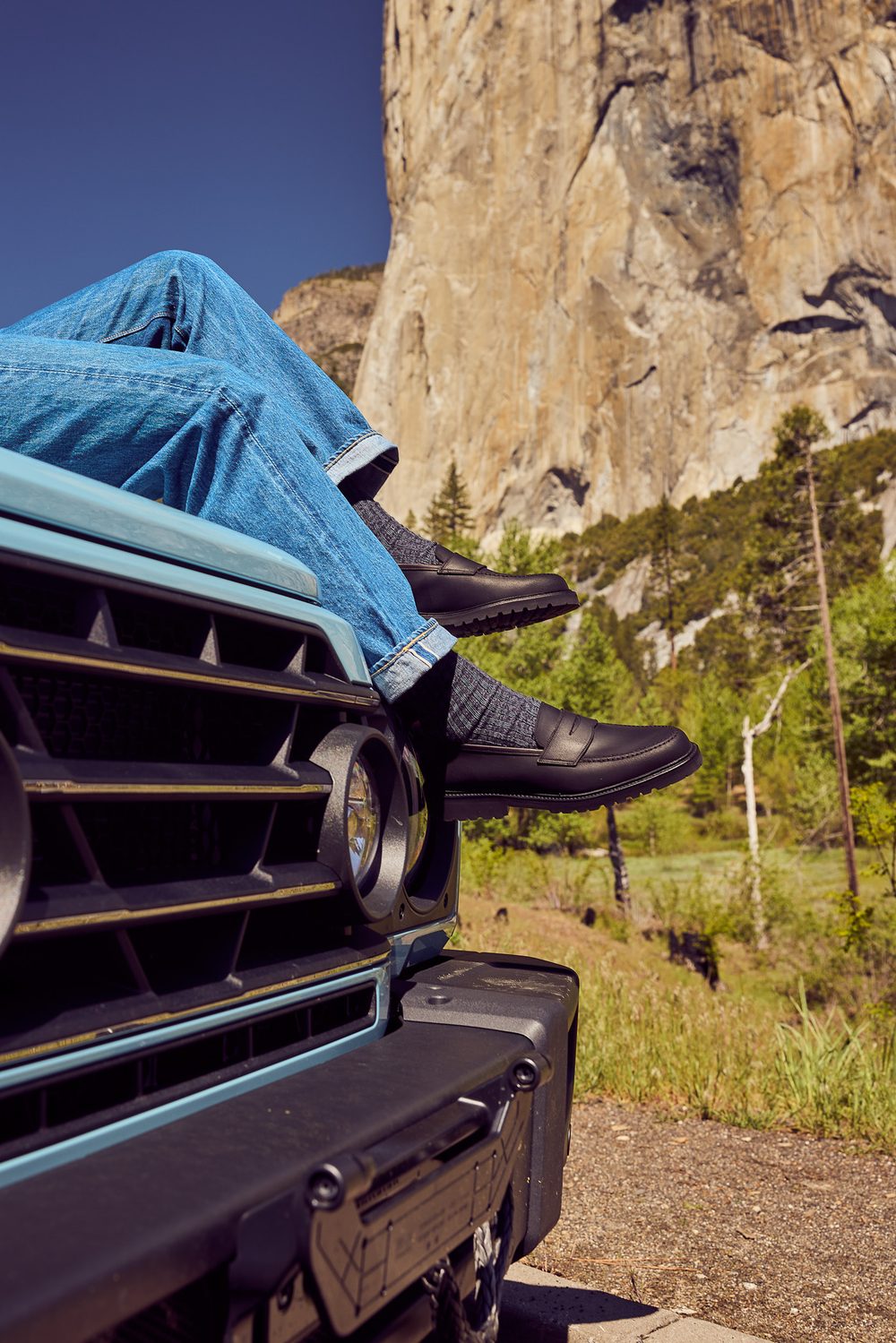 A person wearing blue jeans and dark loafers rests their feet on a light blue vehicle's front.