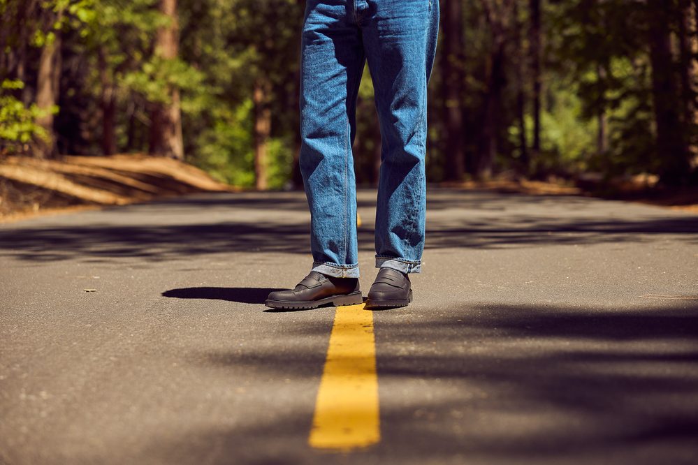 A person stands on a road wearing blue jeans and black shoes with a textured sole.