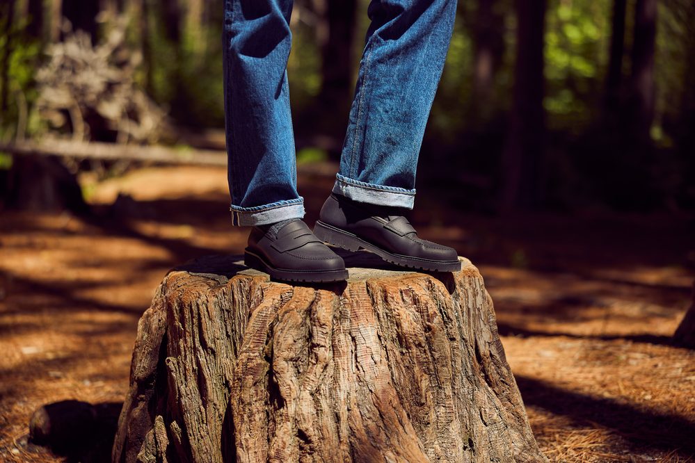 A person stands on a tree stump wearing dark, rugged shoes and blue jeans with rolled cuffs.