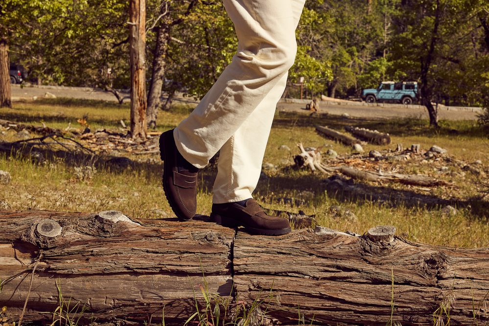 A person wearing cream-colored pants and dark brown loafers is stepping on a log.
