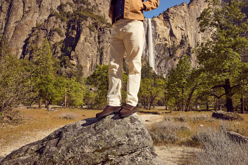 A person stands on a rock wearing brown shoes, cream-colored pants, and a tan jacket.