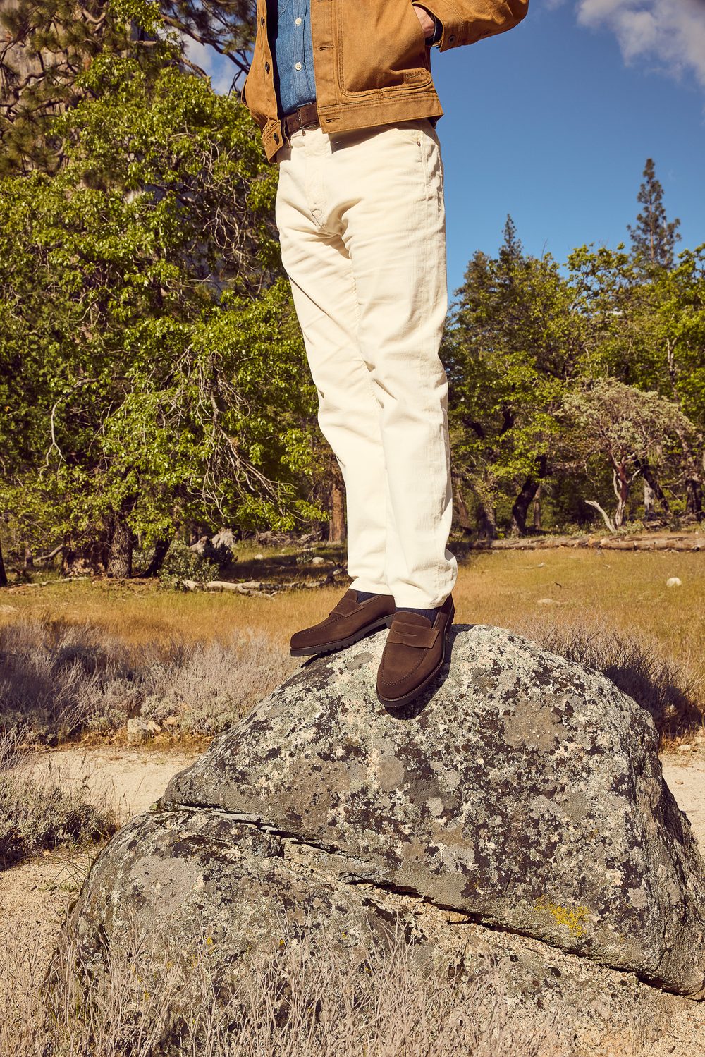A person stands on a large rock wearing brown suede shoes, cream-colored pants, and a brown jacket over a denim shirt.