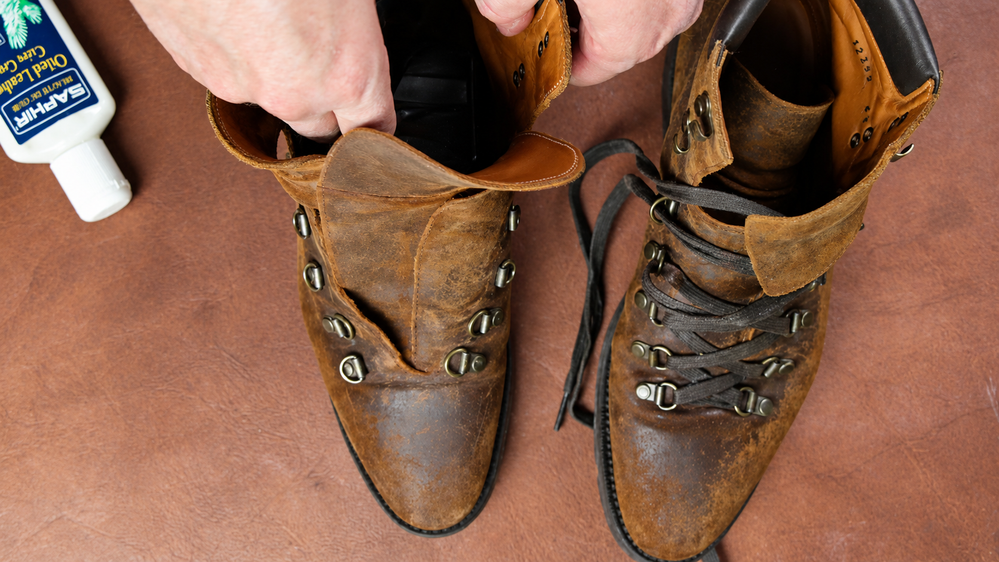 A pair of worn brown leather boots with metal eyelets, one being held open while Saphir Oiled Leather Cream is positioned nearby.