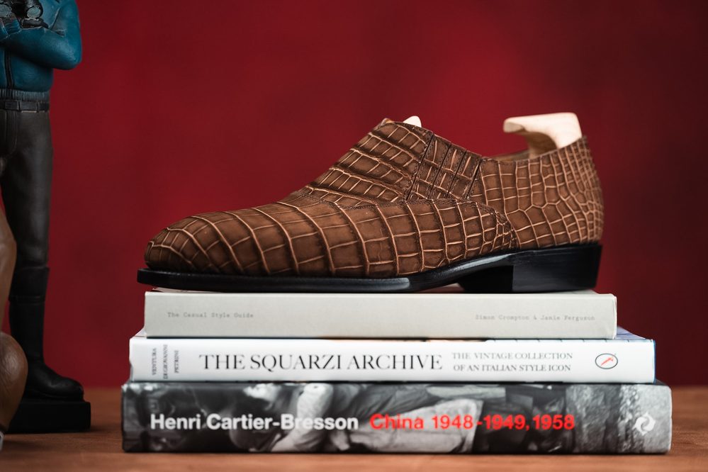 A brown crocodile-embossed leather shoe is displayed on a stack of three books.