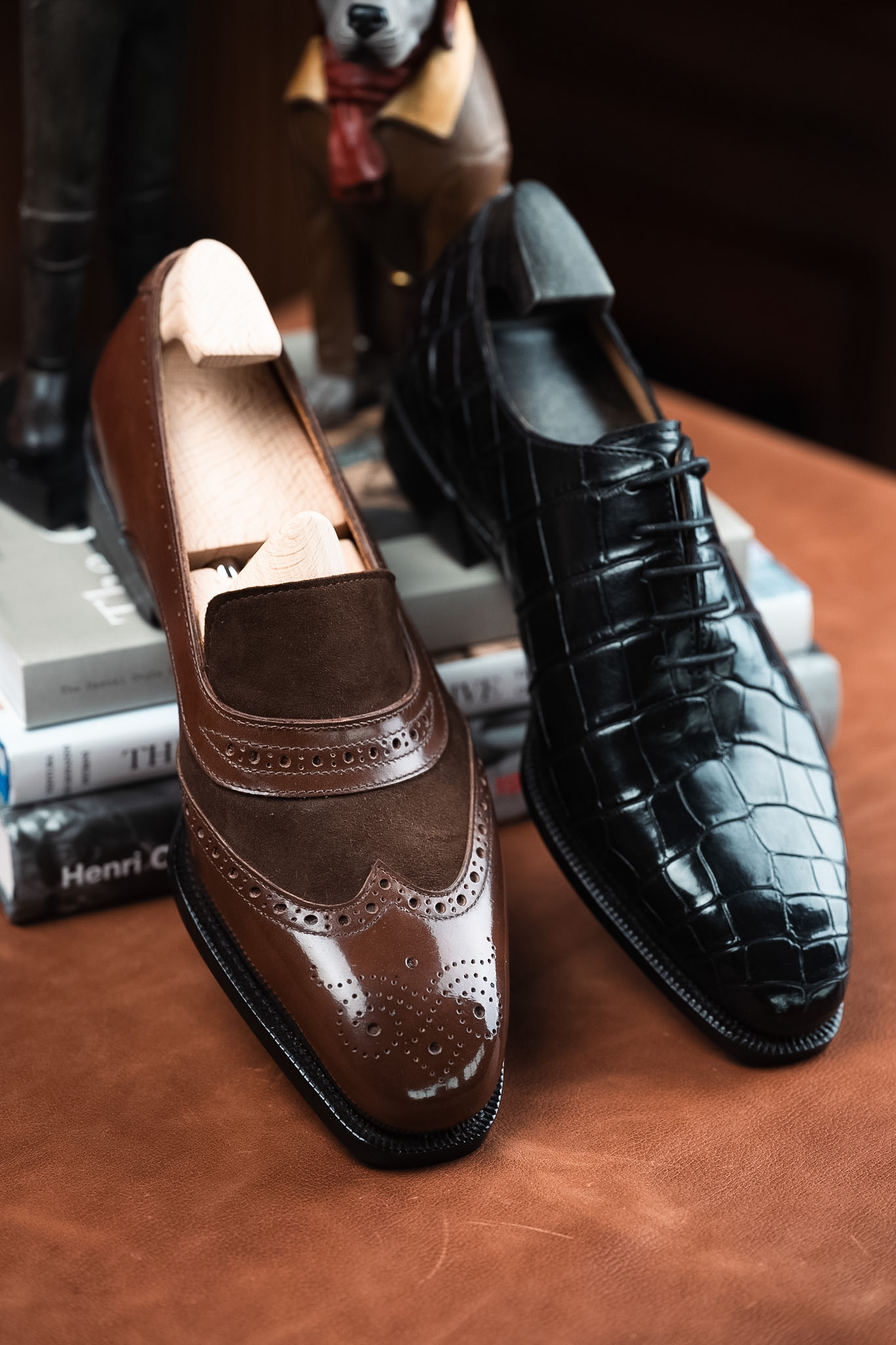 A brown leather shoe with a suede panel and decorative perforations is paired with a black crocodile-patterned leather shoe.