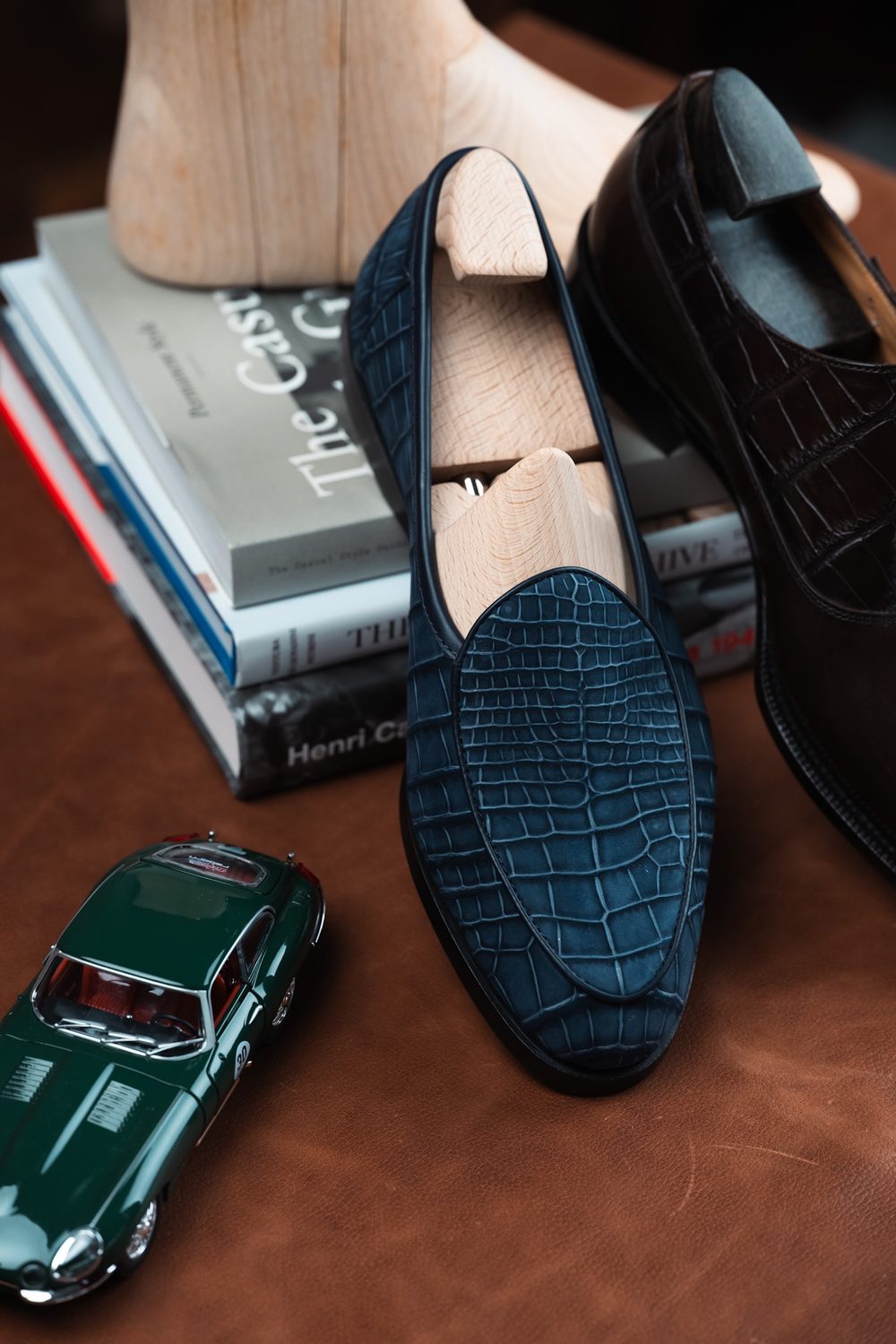 A dark blue crocodile nubuck shoe is displayed alongside a wooden shoe tree and a green toy car, resting on a stack of books.