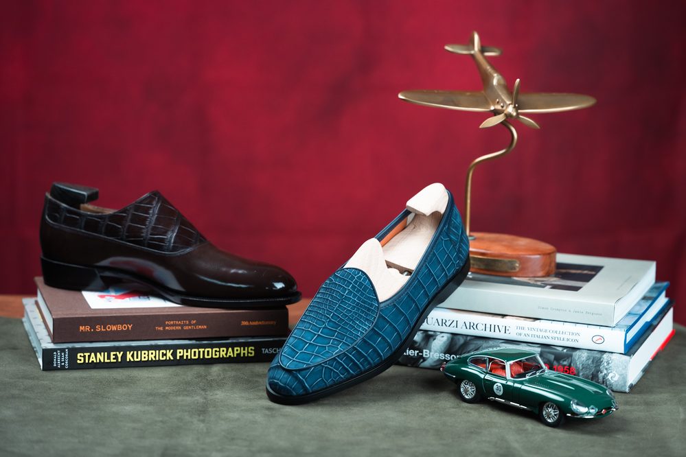 A brown leather shoe and a blue crocodile nubuck loafer are displayed on stacked books.
