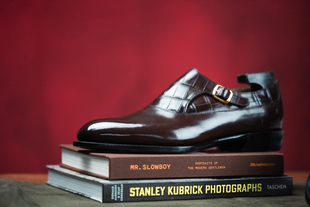 A polished brown leather shoe with a crocodile texture and gold buckle, resting on stacked books.