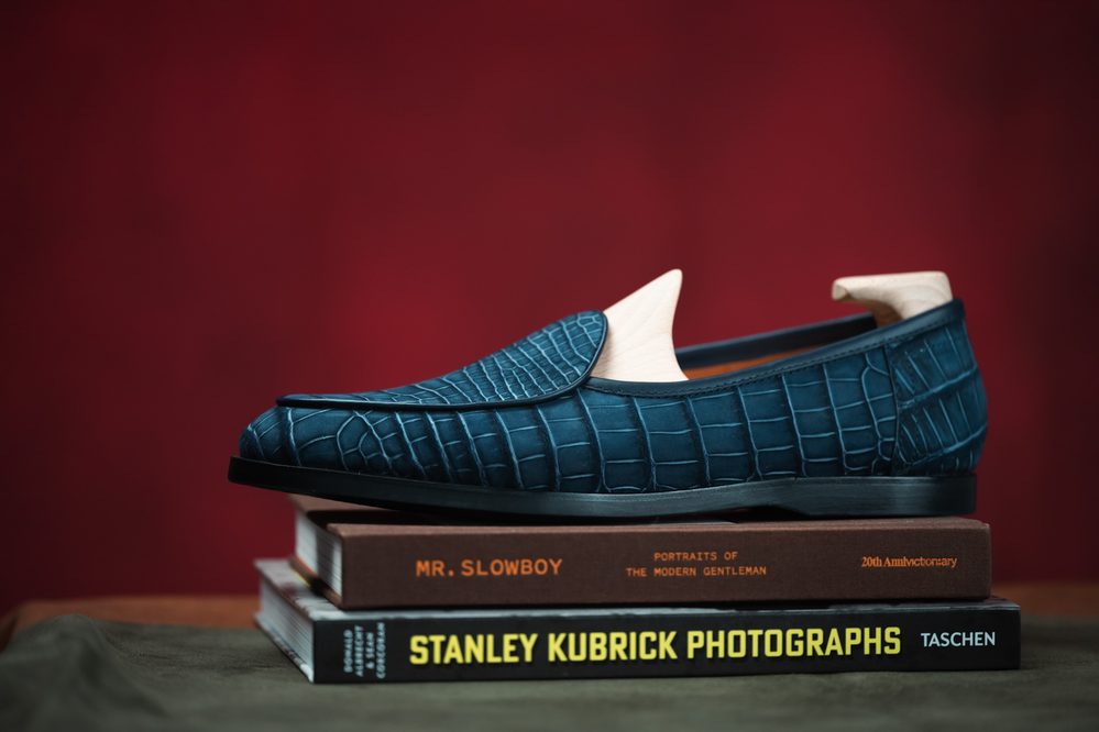 A blue crocodile nubuck shoe with a wooden shoe tree rests on a stack of books.