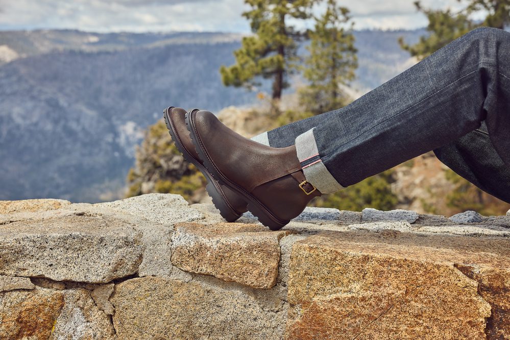 A brown leather boot with a buckle rests on a stone ledge, paired with rolled-up denim jeans.