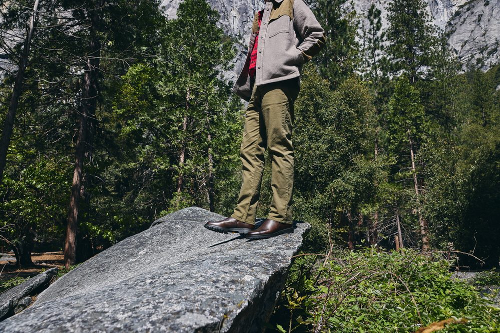 A person stands on a large rock, wearing olive green pants and brown leather shoes, with a gray jacket and red shirt.
