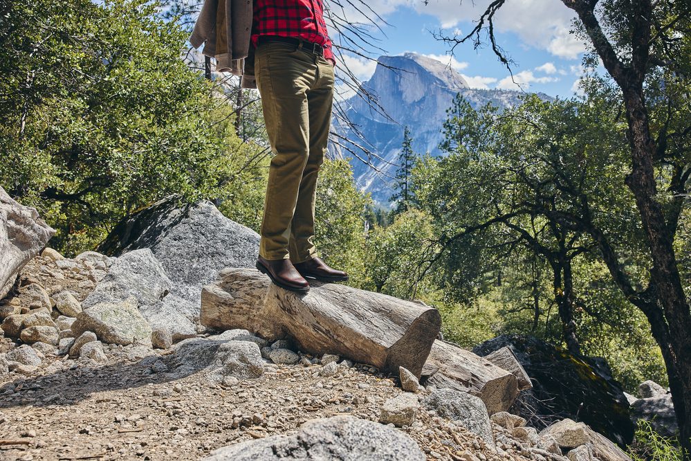 A person stands on a log wearing brown leather boots, olive green pants, and a red plaid shirt.