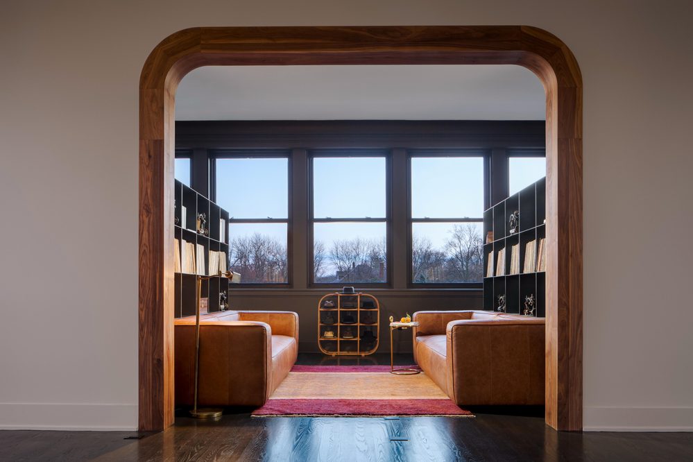 A cozy seating area features two brown leather sofas, a small round table, and a black bookshelf filled with items, framed by a wooden arch.