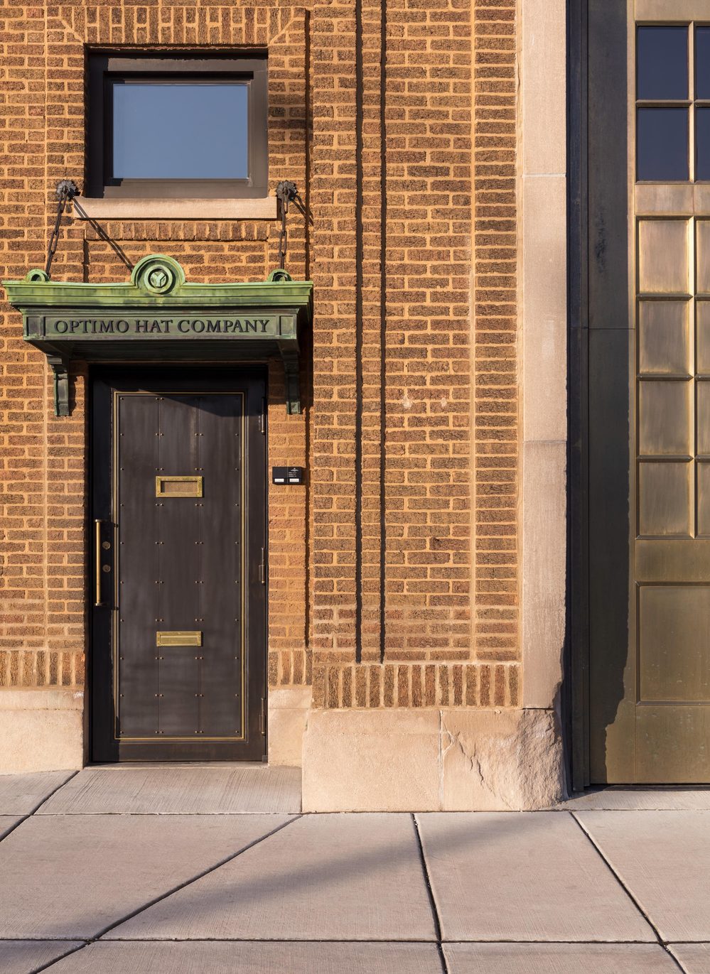 A black door with a brass handle and a green awning labeled "Optimo Hat Company" on a brick wall.
