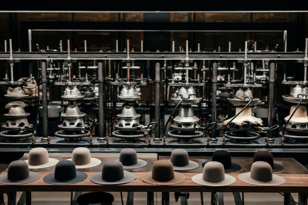 A variety of felt hats in shades of beige, gray, and black are displayed on a wooden table, with hat-making equipment visible in the background.