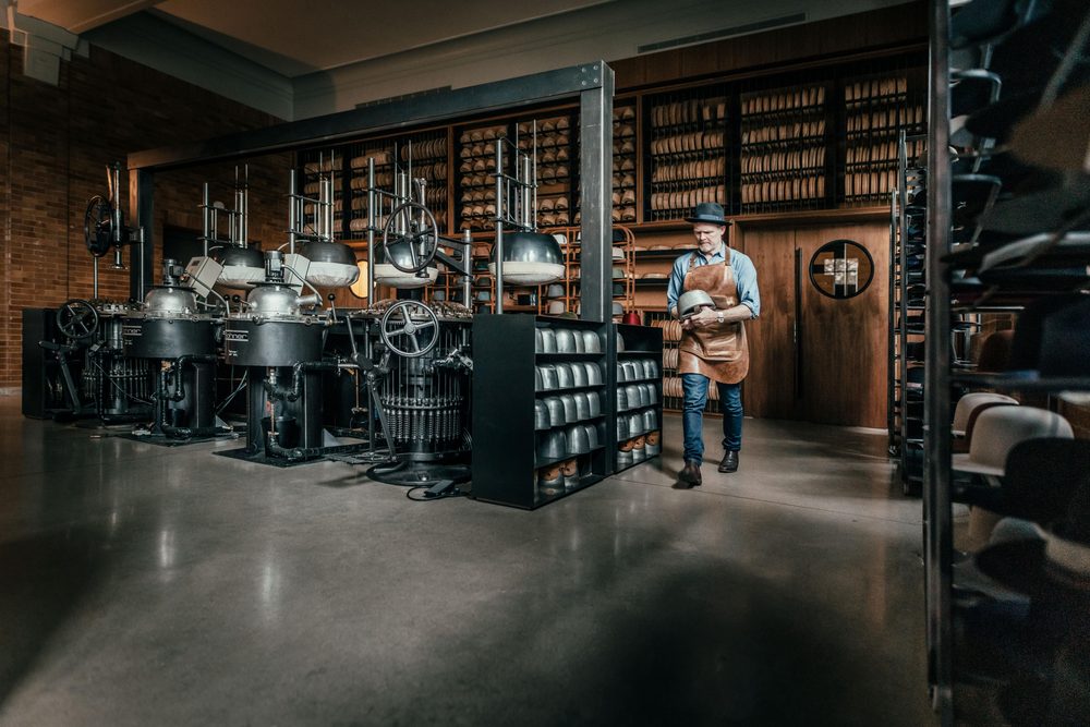 A man wearing an apron and a hat holds a hat while standing near vintage black hat-making machines.
