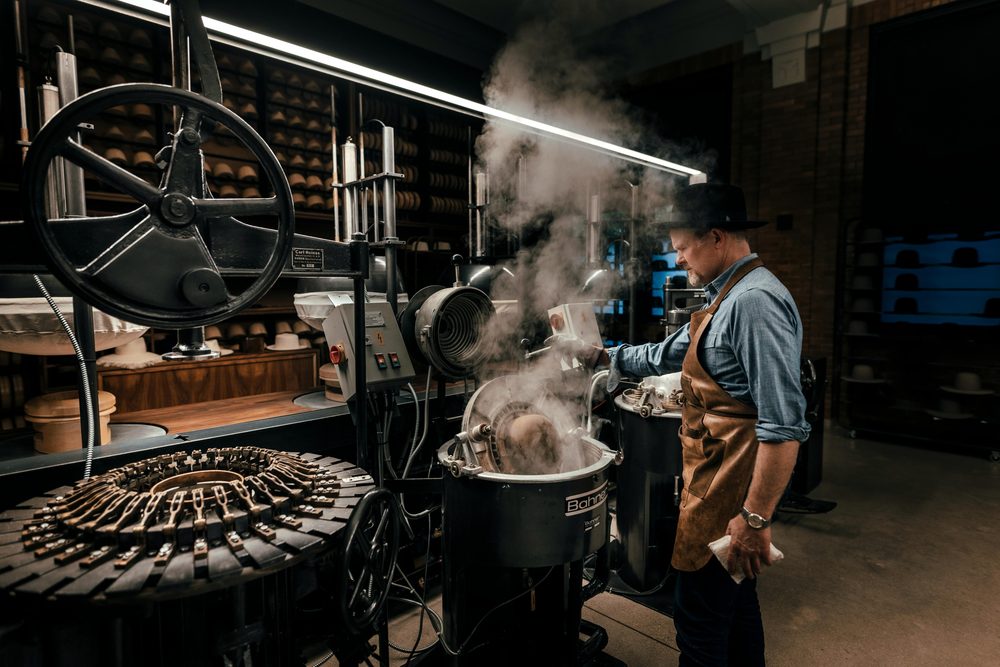A man in a denim shirt and leather apron operates a steam machine at a hat factory.
