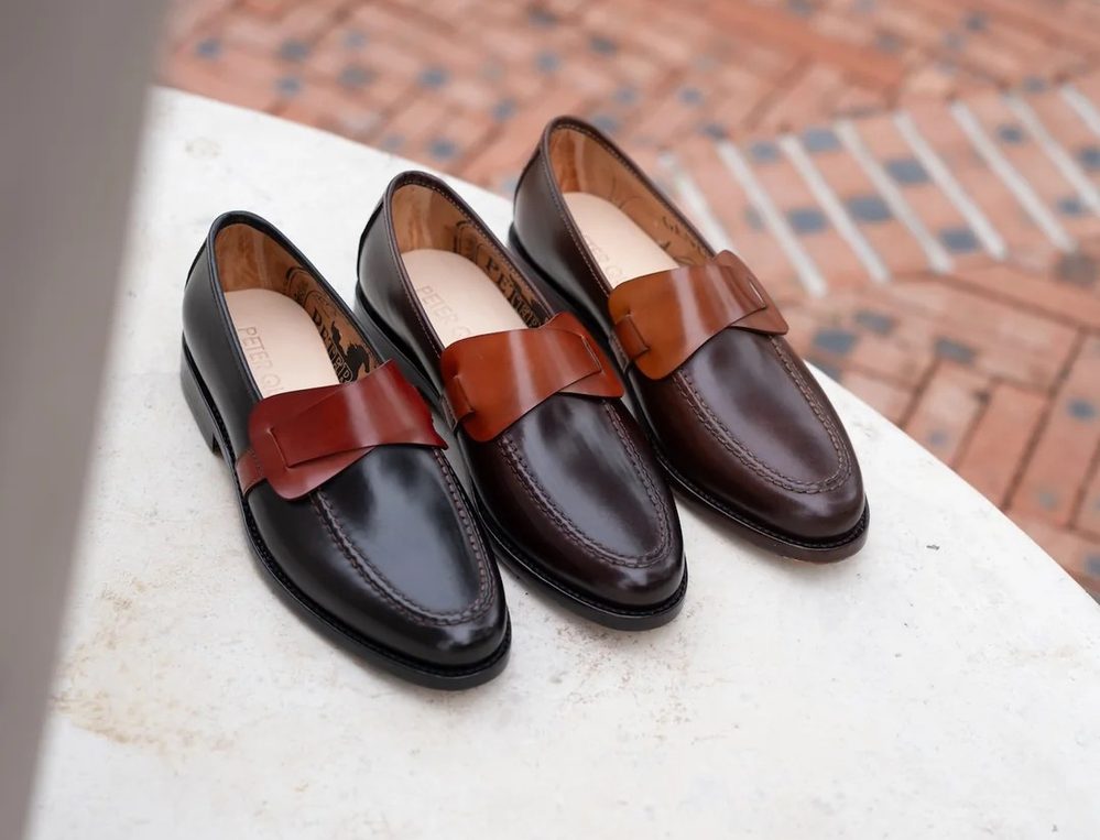 Two pairs of cordovan loafers in black and dark brown leather, each featuring a contrasting brown leather bow.