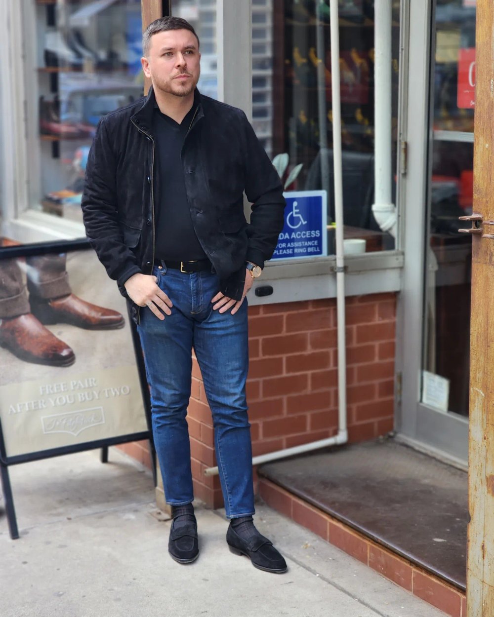 A man wearing a black suede jacket and black suede shoes stands casually, paired with blue jeans and a black shirt.