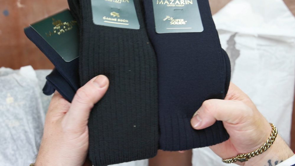 Two pairs of socks in black and navy blue are being held, each with a label showing the brand "MAZARIN."