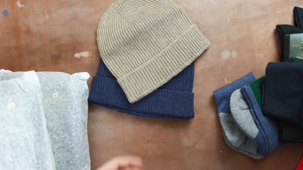 Two knitted hats, one beige and one navy, alongside three pairs of socks in gray, blue, and dark colors, with some wrapped items in tissue paper.