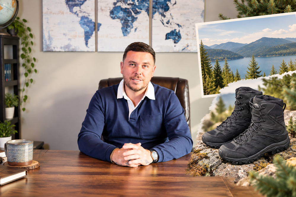 A man in a navy blue shirt sits at a wooden desk, with black hiking boots and an outdoor landscape image displayed beside him.