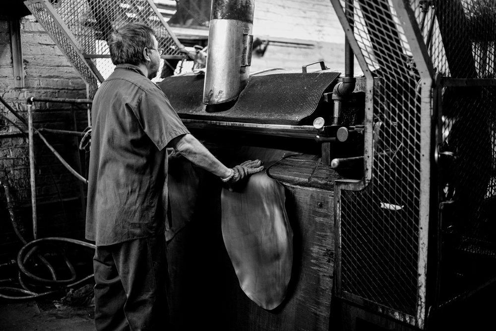 A craftsman operates a large leather pressing machine while handling a piece of leather in a workshop for the Carmina Bespoke Trunk Show.