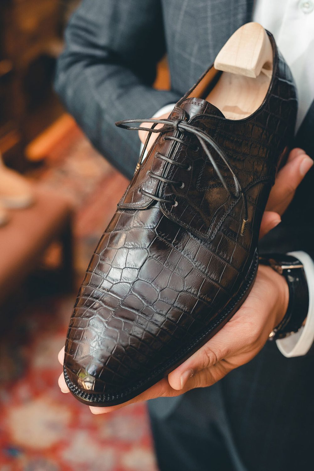 A person holds a dark brown, crocodile-patterned leather shoe with a wooden shoe tree inside.