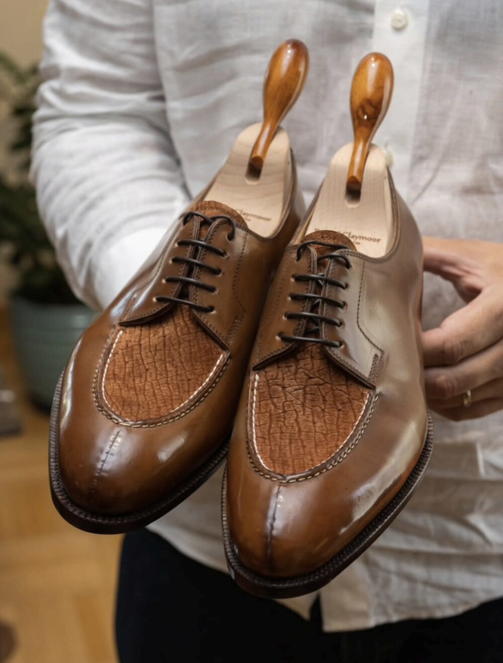 A pair of handmade brown leather shoes featuring a textured, lighter brown panel and wooden shoe trees.