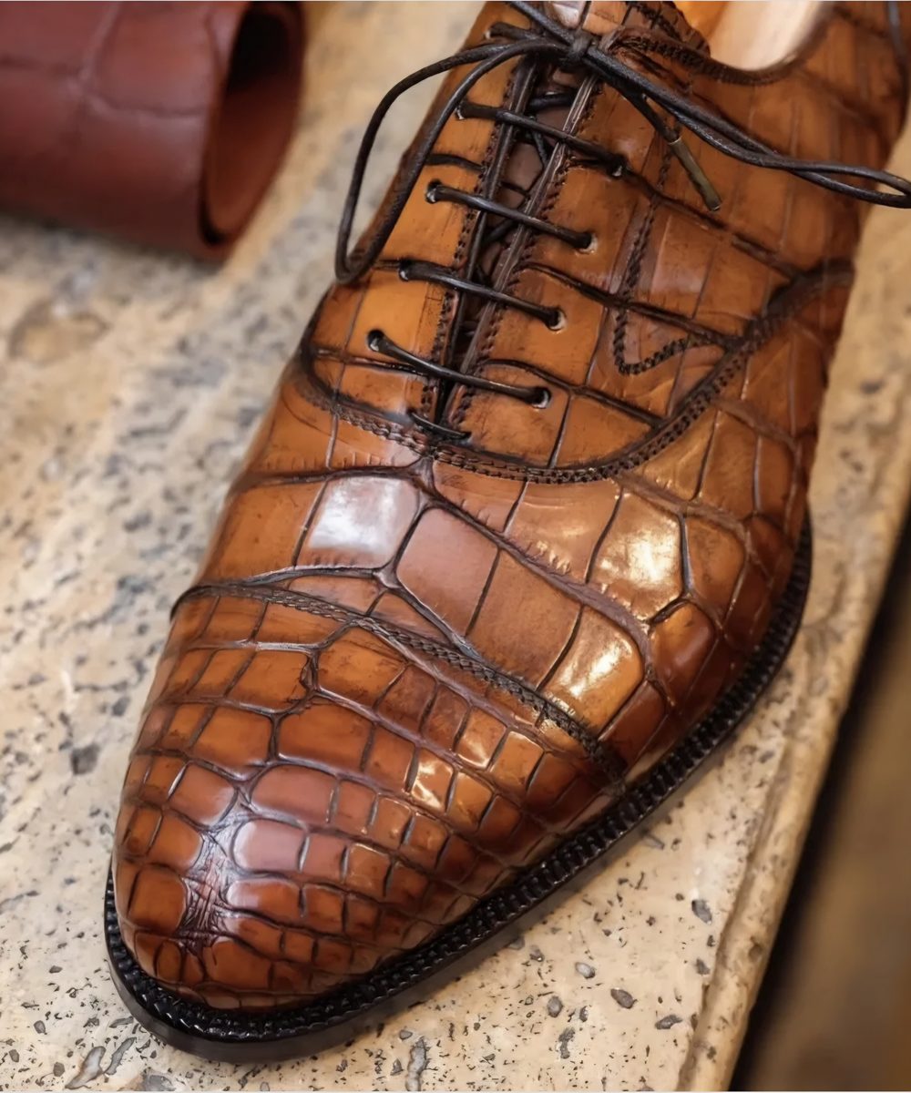 A close-up of a handmade brown leather shoe with a crocodile texture and black laces.
