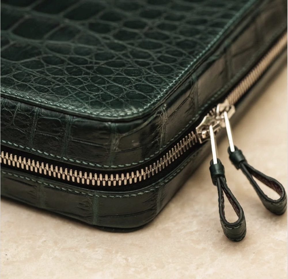 A close-up of a dark green, crocodile-embossed leather bag with a silver zipper and leather pull tags.