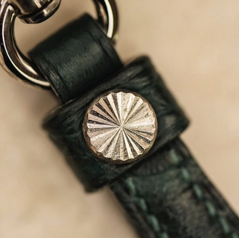 A close-up of a silver metal button with a textured design on a dark green leather strap.