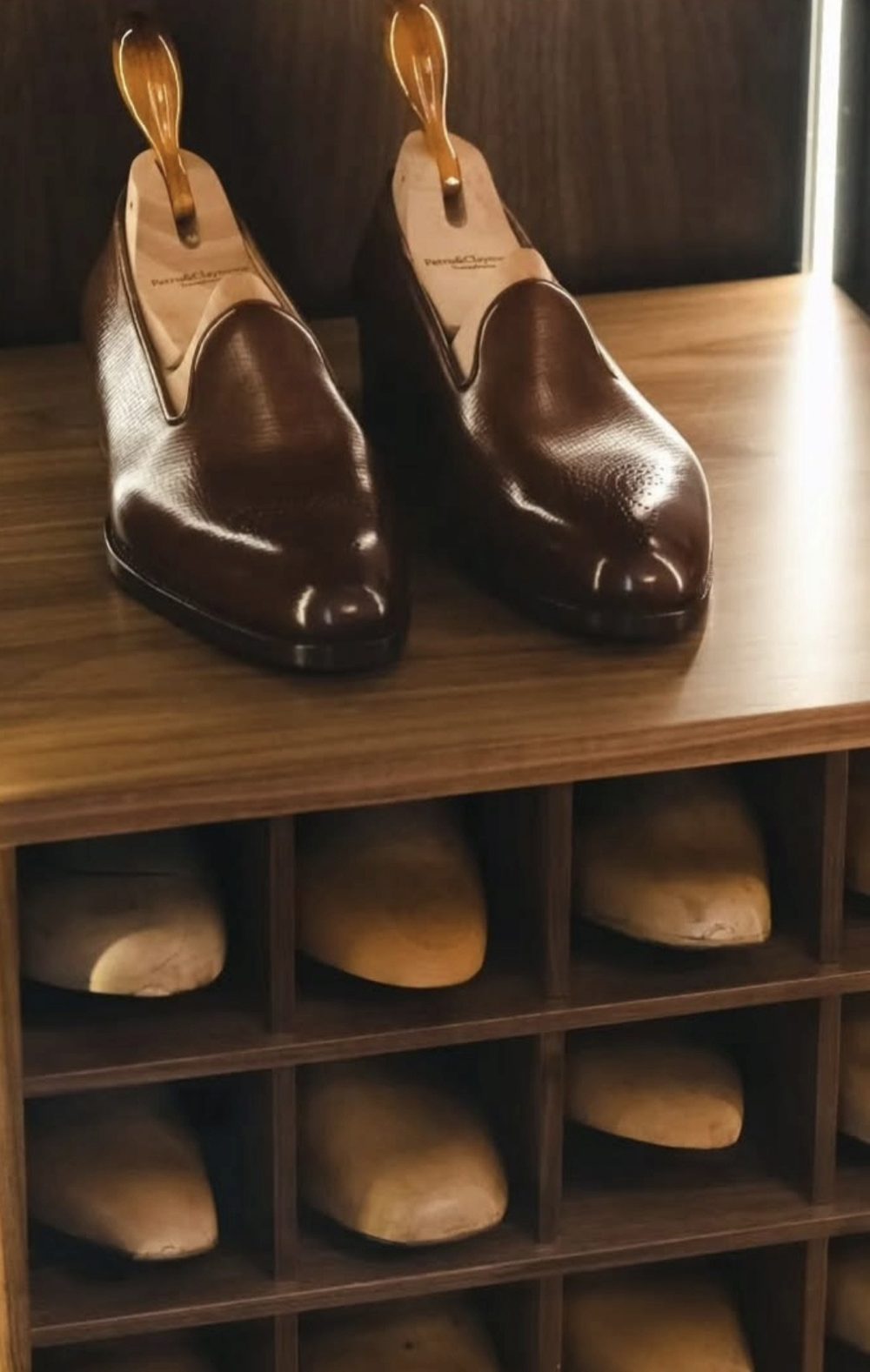A pair of brown handmade leather shoes with wooden shoe trees displayed on a wooden shelf.