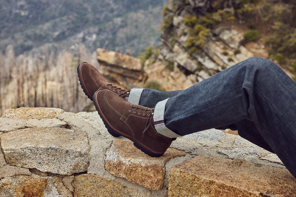 A pair of brown suede boots with white stitching, worn with dark denim jeans, resting on a stone surface.