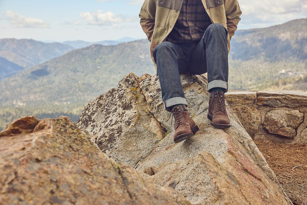 A person wearing brown hiking boots, dark jeans, and a green jacket sits on a rock.
