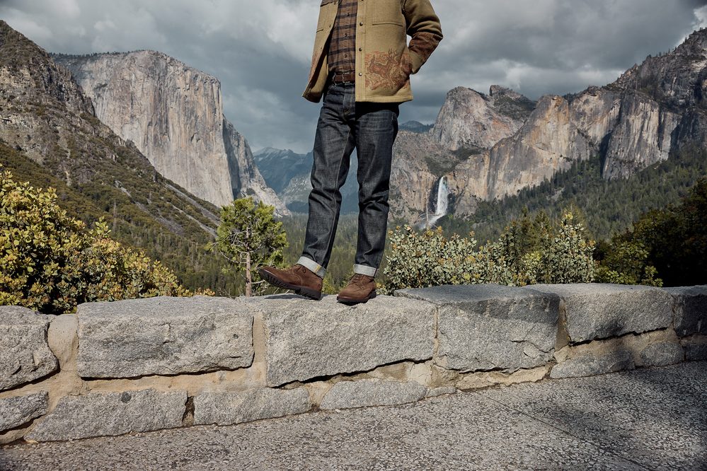 A person stands on a stone wall wearing brown shoes, dark jeans, and a green jacket with an embroidered design.