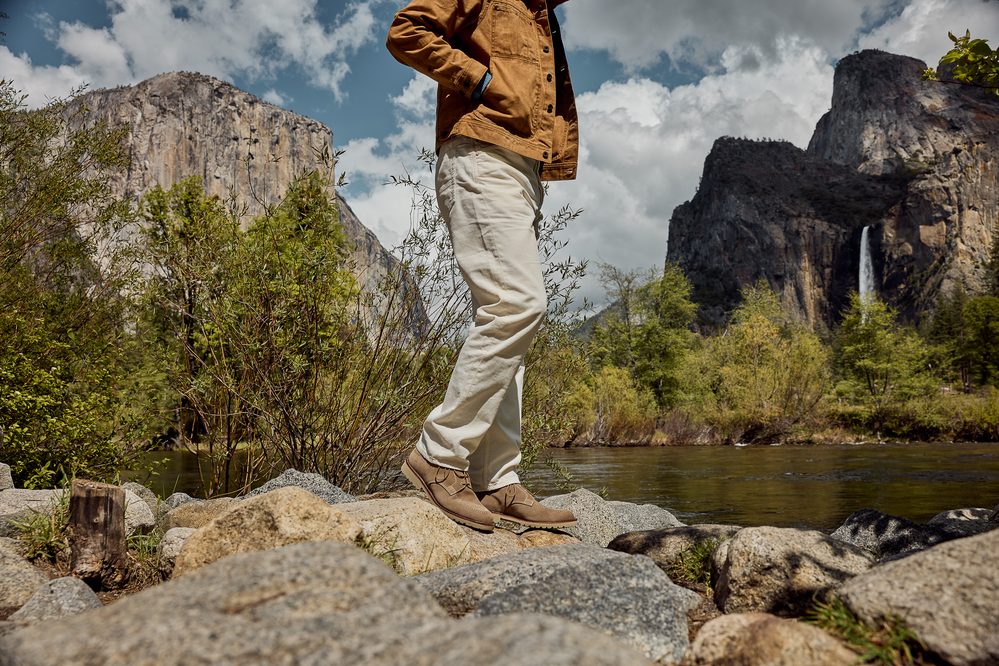A person stands on rocks by a river, wearing tan shoes and light-colored pants with a brown jacket.
