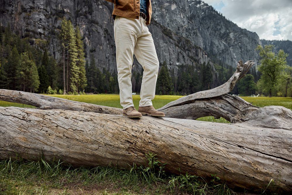 A person stands on a large log wearing beige shoes and light-colored pants with a brown jacket.