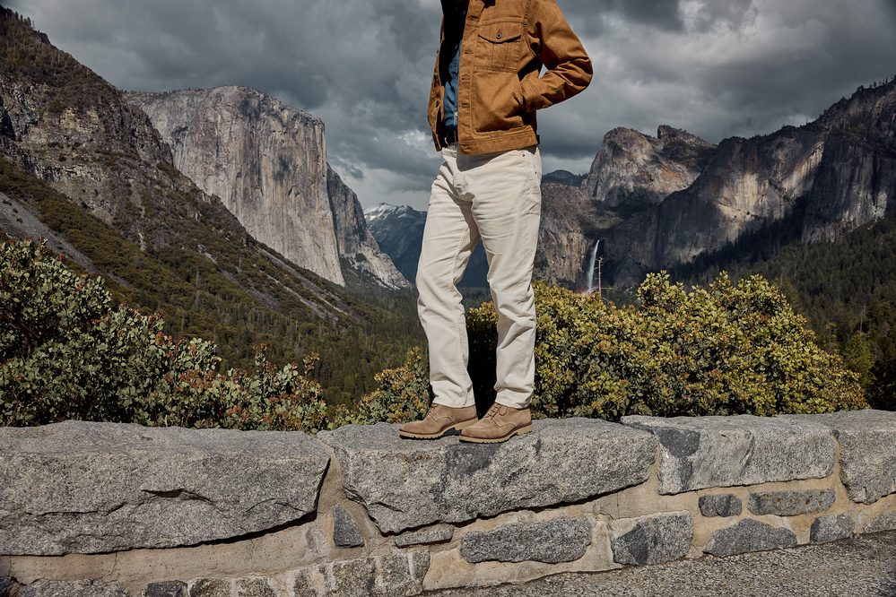 A person stands on a stone wall wearing a brown jacket, white pants, and tan boots.